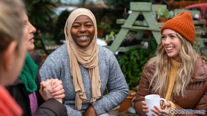 Volunteers sitting outdoors wearing warm casual clothing on a sunny cold winters day. They are resting and having a tea break from working on a community farm, looking after crops and performing other sustainable and environmentally friendly tasks. They are laughing and talking together, drinking hot drinks.